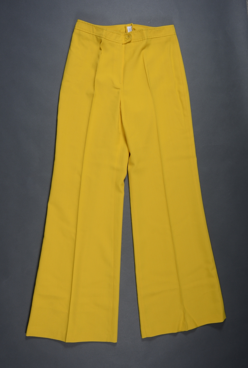 Slengbukse til dame sydd av gul terylene. Det er en enkel modell. I livet er det linning med knapp og gylflukning. Buksen har ikke lommer. På beina er det presser. Buksebeina har bra vidde nederst.