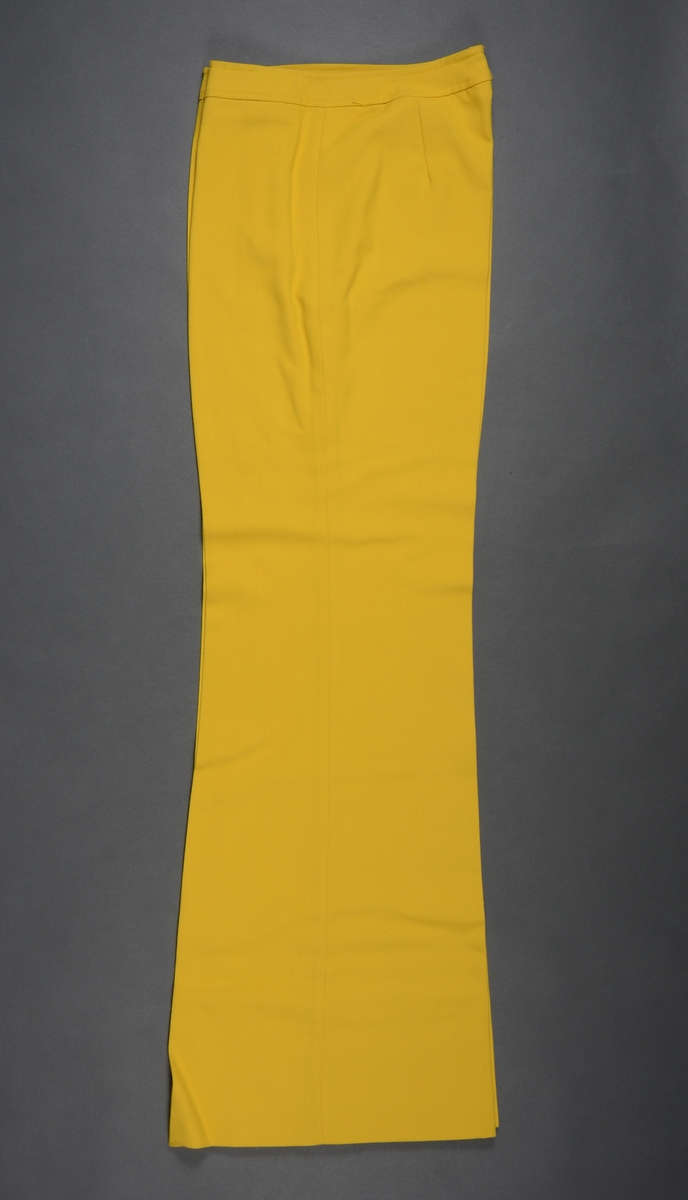 Slengbukse til dame sydd av gul terylene. Det er en enkel modell. I livet er det linning med knapp og gylflukning. Buksen har ikke lommer. På beina er det presser. Buksebeina har bra vidde nederst.