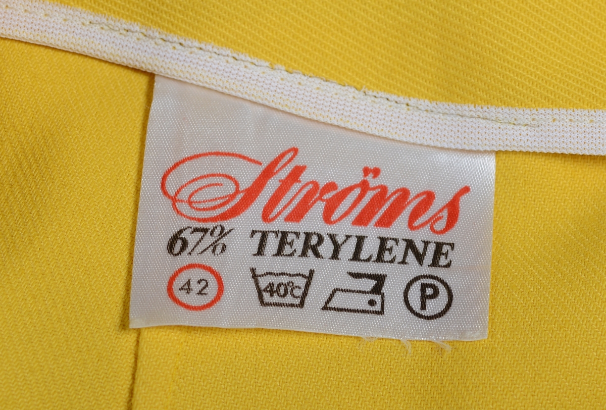 Slengbukse til dame sydd av gul terylene. Det er en enkel modell. I livet er det linning med knapp og gylflukning. Buksen har ikke lommer. På beina er det presser. Buksebeina har bra vidde nederst.