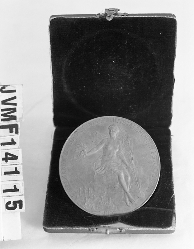 Minnesmynt från Stockholmsutställningen 1897.
Myntet är av brons, och visar på ena sidan en profilbild av Oscar II i halvrelief. På den andra sidan syns en halvnaken kvinnofigur hållande ett palmblad (?) i ena handen och en lagerkrans i den andra handen.
Text på ena sidan: "Oscar II Sveriges och Norges konung - Brödrafolkens väl".
Text på andra sidan: "Allmänna konst och industriutställningen i Stockholm 1897".

Myntet förvaras i en tillhörande ask fodrad med brun sammet.