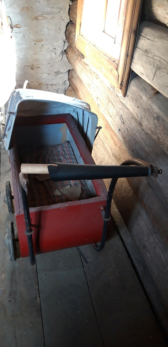 Rødmalt vogn med gråmalt innerside og lys grå kalesje av kunstlær. 4 metallhjul med nedslitt gummidekk. Håndtak av tre med svart plastlignende materiale som er sprøtt. Kalesje med ei flenge. Polstret karm. Mangler skrue i håndtak og kalesje.
