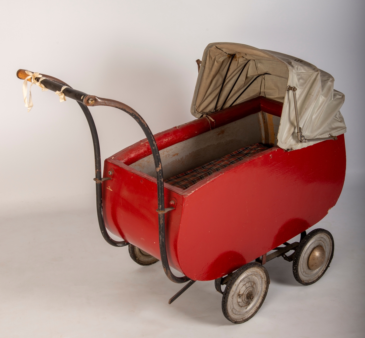 Rødmalt vogn med gråmalt innerside og lys grå kalesje av kunstlær. 4 metallhjul med nedslitt gummidekk. Håndtak av tre med svart plastlignende materiale som er sprøtt. Kalesje med ei flenge. Polstret karm. Mangler skrue i håndtak og kalesje.