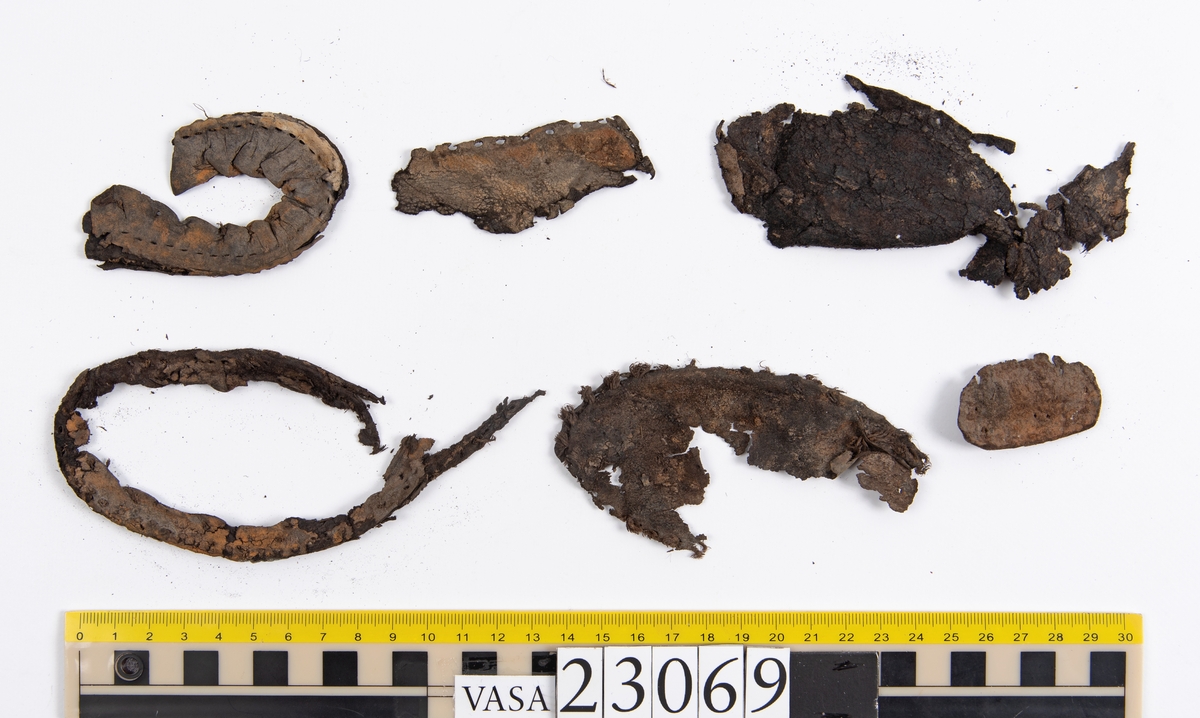 Dokumentation 1996:
Delar och fragment av sko: en skobotten samt ett fragmentariskt ovanläder, tre bitar av ett filtliknande material, ett 15-tal fragment av bottenläder samt sex mindre fragment av trä.
Skobottnen har kvarsittande tråd och träpligg. Yttersulan är ritsad. Klacken består av fem klacklappar som är placerade mellan ytter- och innersulan. Innersulans läder är mycket kraftigt, ca 6 mm tjock. Ovanlädret är sprött och fragmentariskt samt har blå och rostfärgade utfällningar.

Text in English: 
Various parts and fragments of a shoe, consisting of a sole with fragments of vamp (upper leather), three pieces of a felt-like material, some 15 fragments of sole or bottom leather and six smaller fragments of wood.
There are remains of thread and wooden pegs on the shoe sole. The outsole is channeled. The heel is made up of five heel lifts placed between the insole and outsole. The leather of the insole is very thick, ca 6 mm. The upper leather is fragile and fragmentary, and has blue and rust-coloured precipitations.

Dokumentation 2022:
Skobotten och delar av ovanläder till en öppen randsko. Tillverkade i getskinn av god kvalitet. Skapades på en rak läst och bars på höger fot. Klacken som är lagad med en läderlapp under sulan. Inga dekorationer. Det finns tecken på delaminering. Fragment från en stickad strumpa hittades inuti skon. Det finns lagningar med träpligg på sulan och hälen. Skobotten hör ihop med fyndnummer 23070.
För mer information, se Länkade filer: Uppritning

Text in english:
This is the bottom unit and upper fragments of an open shoe, made of goatskin leather of good quality, of randed construction with a patch heel, made on a straight last and worn on the right foot, with no decorative features. The bottom unit pairs with 23070. There are signs of delamination and fragments of a knitted stocking were found inside the shoe. There are pegged repairs to sole and heel.
For more information, go to Länkade filer: Uppritning