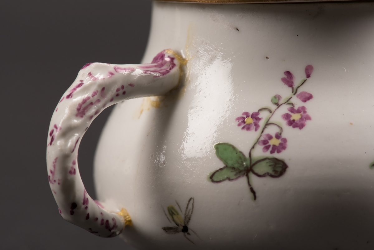 Krämkopp med öra och lock tillverkad av fajans. Bemålad med blommor, frukter och insekter i flera färger på vit botten. Lockknoppen är utformad som en frukt med skaft och blad i relief.