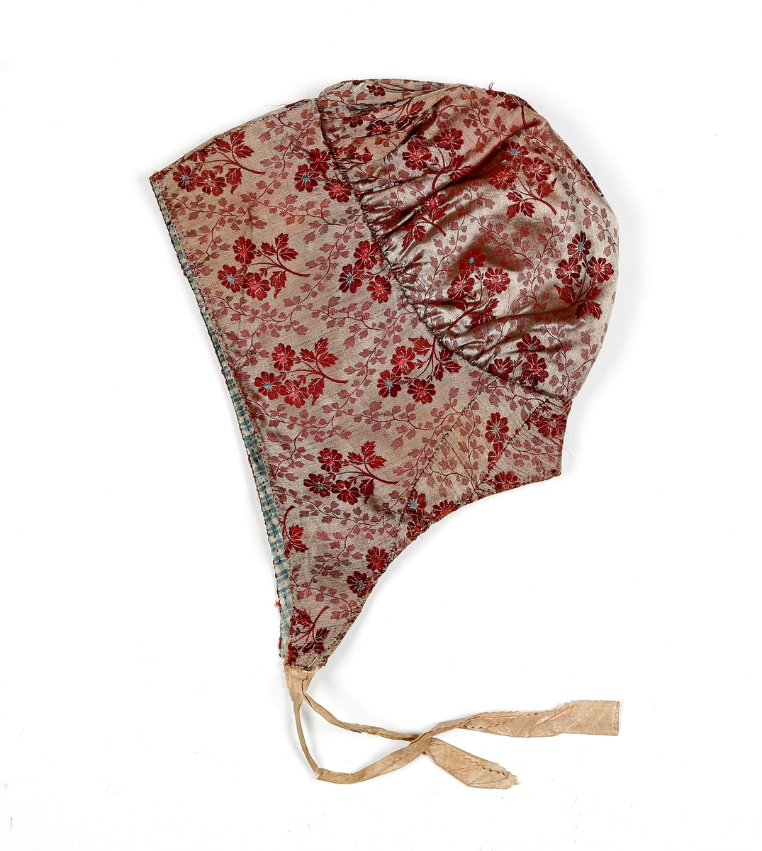 Høglue i mønstret silke med beige knytebånd som er sikk-sakk-klippet i kantene. Lua er fòret med smårutete bomullsstoff.  Pannestykket er sydd i to hoveddeler der det er skjøtt inn to stykker i den ene halvdelen. Rundt kanten er det sydd kastesting med prikk. Pullen er rynket til pannestykket.