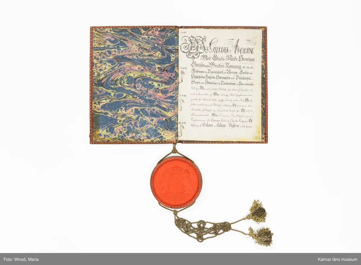 KLM 16024. Sköldebrev, adelsbrev, läder, papper, färg, metall, textil. Sju sidor med handskrift på pergament, inbunden med gul och grön tvinnad snodd med tofsar. På sida tre kolorerad bild av adelsvapnet. Pärm i rött skinn (chagrinband). Guldtryck fram och baksida med Gustaf IV Adolfs krönta monogram samt dekor i guld runt pärmens kanter. Vidhängande lacksigill med svenska riksvapnet i försilvrad kapsel.