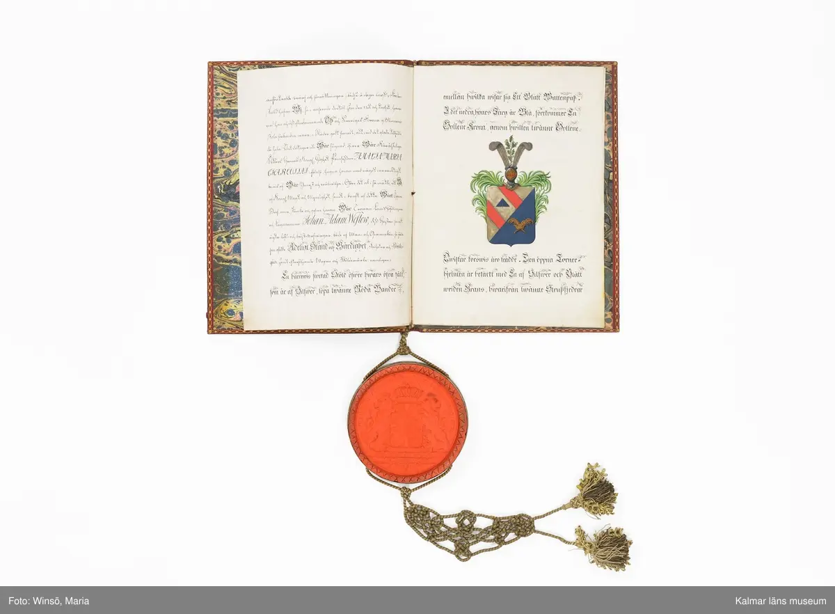 KLM 16024. Sköldebrev, adelsbrev, läder, papper, färg, metall, textil. Sju sidor med handskrift på pergament, inbunden med gul och grön tvinnad snodd med tofsar. På sida tre kolorerad bild av adelsvapnet. Pärm i rött skinn (chagrinband). Guldtryck fram och baksida med Gustaf IV Adolfs krönta monogram samt dekor i guld runt pärmens kanter. Vidhängande lacksigill med svenska riksvapnet i försilvrad kapsel.