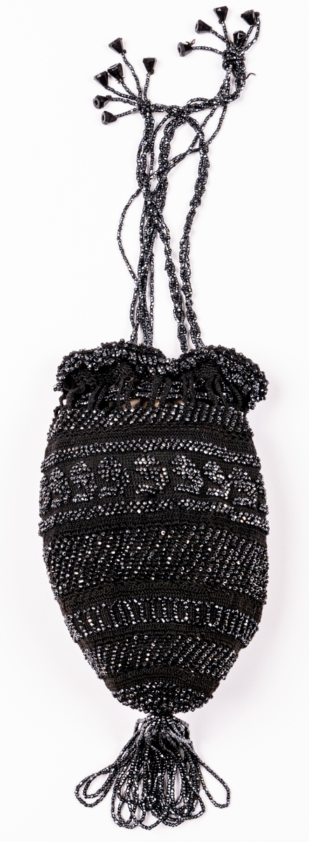 Aftonväska, påsmodell, svart, virkad med inträdda pärlor. Nertill pärlfransar. Väskan bärs i pärlklädda snoddar vilka används för att försluta väskan upptill.