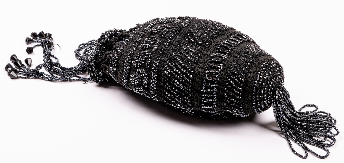 Aftonväska, påsmodell, svart, virkad med inträdda pärlor. Nertill pärlfransar. Väskan bärs i pärlklädda snoddar vilka används för att försluta väskan upptill.