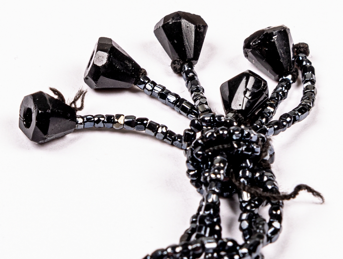 Aftonväska, påsmodell, svart, virkad med inträdda pärlor. Nertill pärlfransar. Väskan bärs i pärlklädda snoddar vilka används för att försluta väskan upptill.