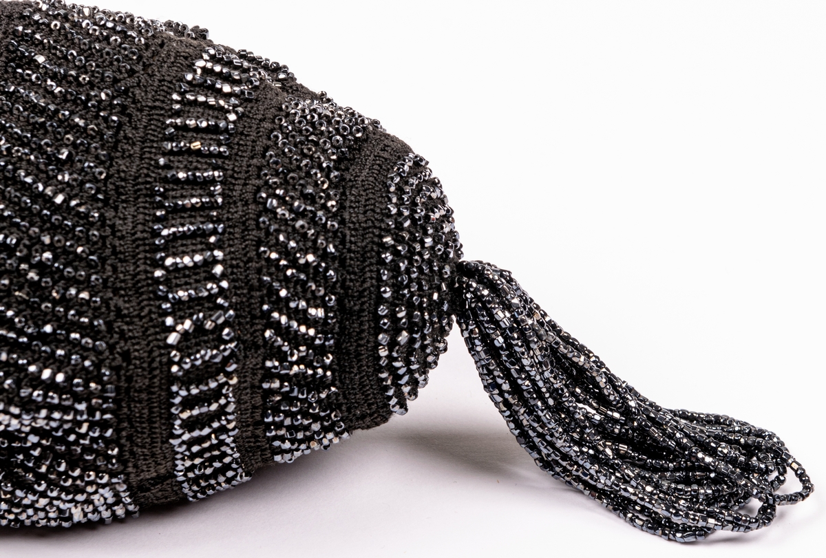 Aftonväska, påsmodell, svart, virkad med inträdda pärlor. Nertill pärlfransar. Väskan bärs i pärlklädda snoddar vilka används för att försluta väskan upptill.