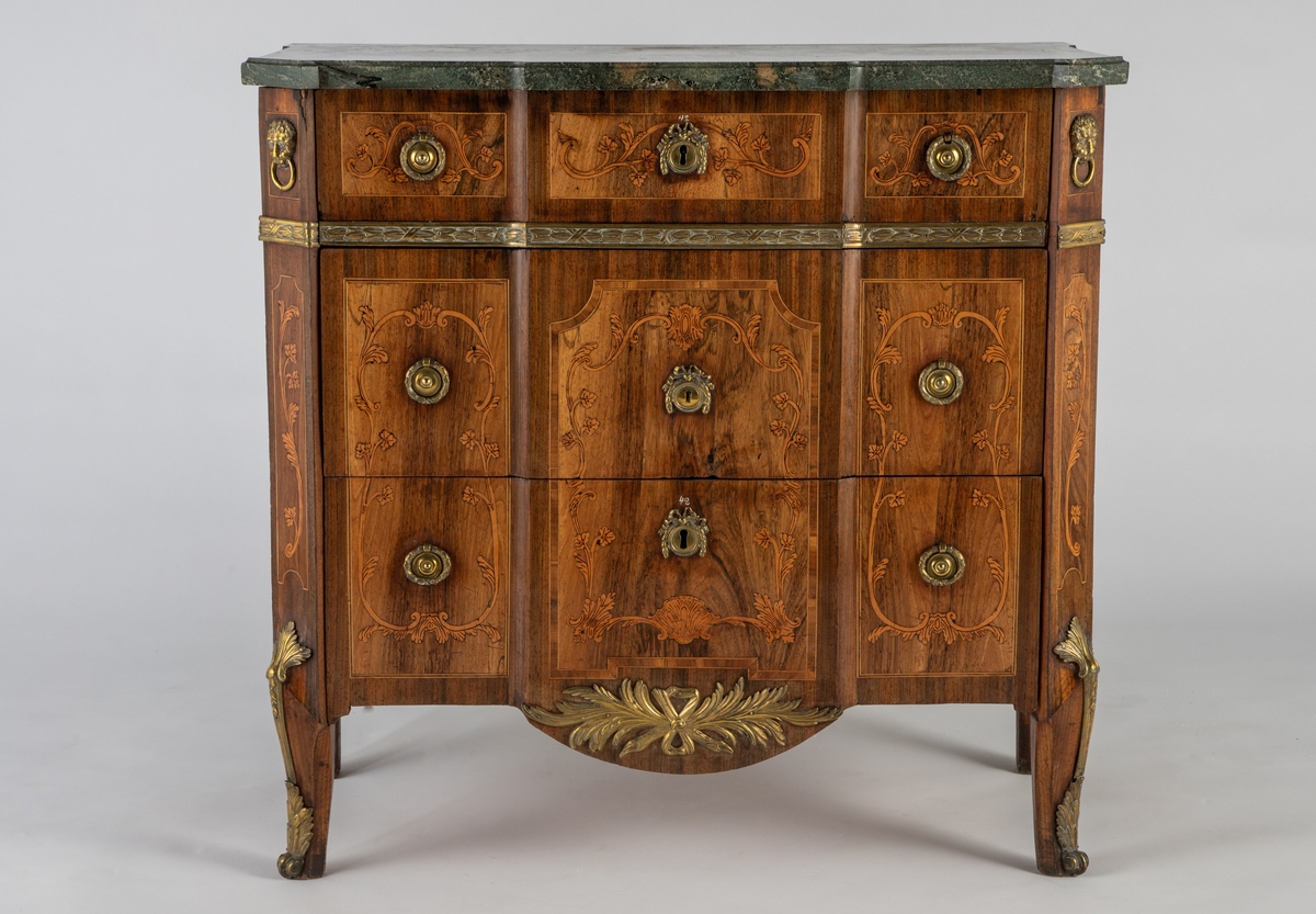 Kommode med knekket front med fremskutt midtparti og avskårne hjørner. Intarsiadekor og bronsebeslag. Profilert marmorplate som følger møbelets form. Forsiden er dekorert med en rekke bronsebeslag: nederst grener med sløyfe, festonger på låsskiltene, rosetter med laurbærkrans på håndtakene, volutter på sidene, palmetter på bena, løvehoder med ring på hjørnene, bronsebånd med laurbærblad under øverste skuff. Intarsiadekor med bladranker.