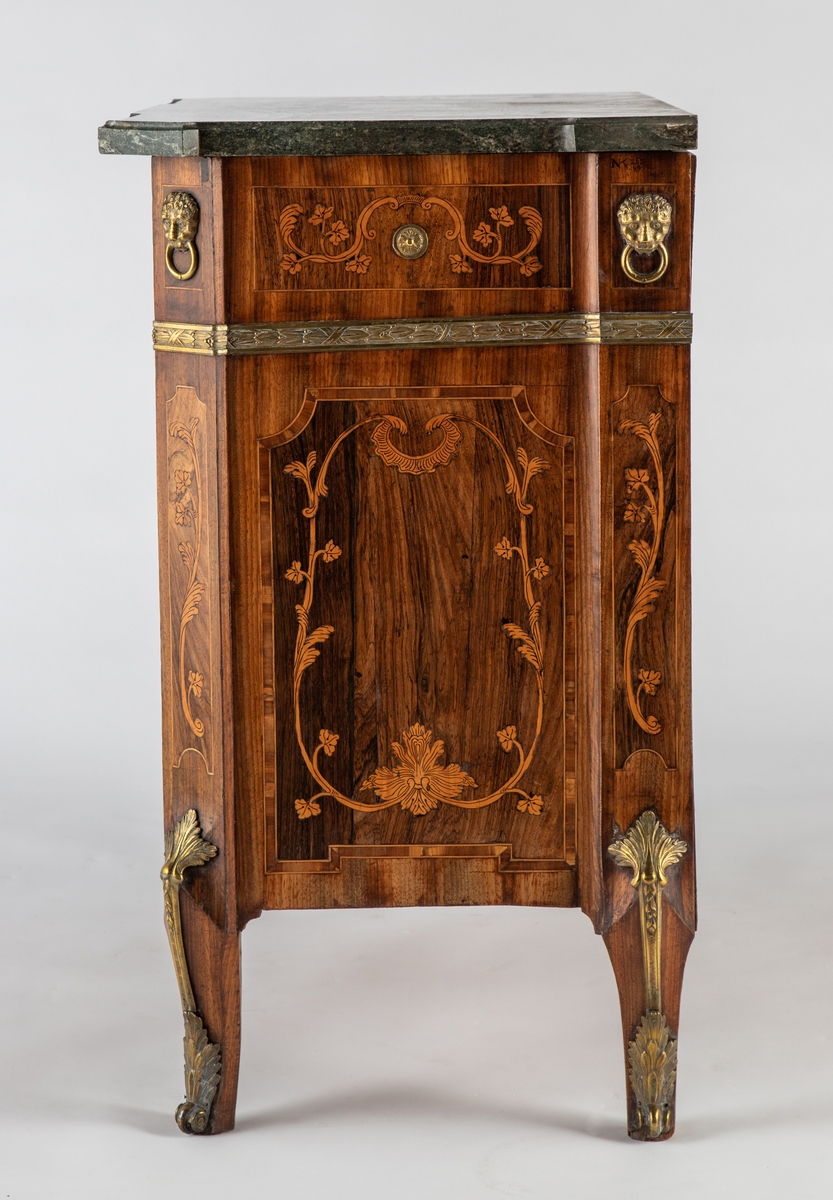 Kommode med knekket front med fremskutt midtparti og avskårne hjørner. Intarsiadekor og bronsebeslag. Profilert marmorplate som følger møbelets form. Forsiden er dekorert med en rekke bronsebeslag: nederst grener med sløyfe, festonger på låsskiltene, rosetter med laurbærkrans på håndtakene, volutter på sidene, palmetter på bena, løvehoder med ring på hjørnene, bronsebånd med laurbærblad under øverste skuff. Intarsiadekor med bladranker.