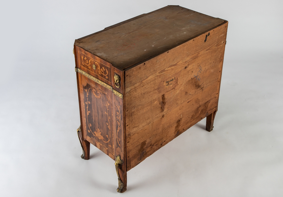 Kommode med knekket front med fremskutt midtparti og avskårne hjørner. Intarsiadekor og bronsebeslag. Profilert marmorplate som følger møbelets form. Forsiden er dekorert med en rekke bronsebeslag: nederst grener med sløyfe, festonger på låsskiltene, rosetter med laurbærkrans på håndtakene, volutter på sidene, palmetter på bena, løvehoder med ring på hjørnene, bronsebånd med laurbærblad under øverste skuff. Intarsiadekor med bladranker.