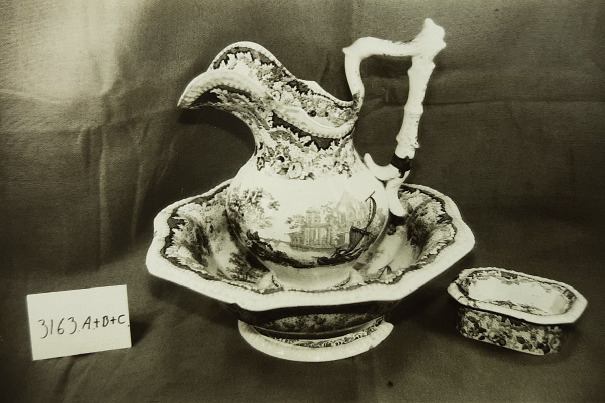 Vaskesett i fajanse, besående av:
A) Vaskevannsfat med sirkulær fotkant med perlebord i relieff. Vid buk med bred blomsterbord. Utbrettet,  8-kantet munningsrand med perlebord langs kanten. Motiv i bunnen: seilbåt med topersoner, hus og bro i bakgrunnen. H: 12.5, Di: 18.5. 

(B) Vaskevannsmugge med sirkulær fotkant, kuleformet buk, sterkt svunget nebb og knekket hank med metallbånd nederst. Dekormotiv i blått, seil med trær og hus på høyre side av buken. Bred blomster-og bladbord innvendig og utvendig ved munningen. H: 29.0, Di:12.5.

(C) Såpekopp, består av to rektangulære 8-kantete deler, begge med blå dekor. Underdelen har blomsterbord langs sidene. Overdelen har gjennomhullet bunn og utoverbrettet munningskant med blå bord. Bunnen har motiv med seilbåt hus og trær. L:12.1, B:8.0, H:5.7. 

(D) Tannbørstebeholder, rektangulær 8-kantet med blå blomsterbord langs sidene. Innvendig to små tugnete tverrvegger til å legge tannbørsten på. L:17.5,B:6.8 H:3.4.