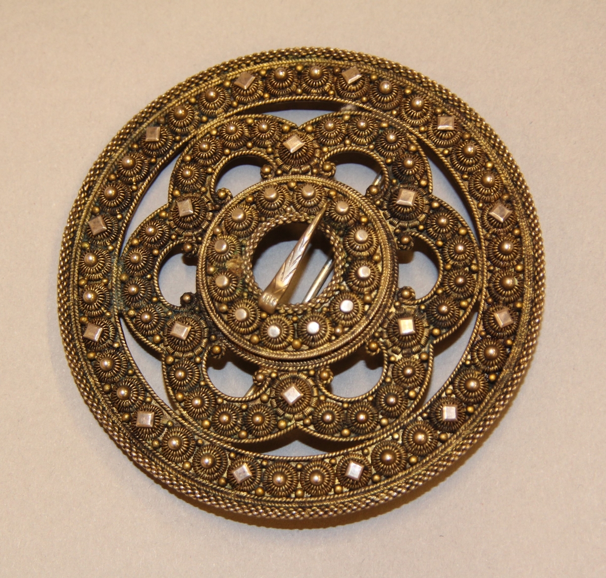 Ein kruse med plate, to krusar med knupp kring ytterste ringen. Inni slangen er det delingskrusar med plater oppå, fire kruser med knupp mellom desse. Rundt spretteholet ei rad med krusar og rund, litt større og flatare knupp. Spretteholet med torn. Sylgja er litt kuva.Nål på baksida.