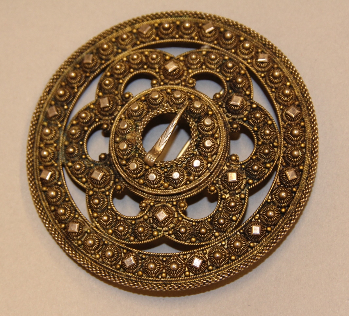 Ein kruse med plate, to krusar med knupp kring ytterste ringen. Inni slangen er det delingskrusar med plater oppå, fire kruser med knupp mellom desse. Rundt spretteholet ei rad med krusar og rund, litt større og flatare knupp. Spretteholet med torn. Sylgja er litt kuva.Nål på baksida.