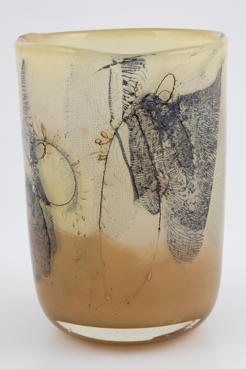Sylinderformet vase i halvgjennomskinnelig glass. Ujevn munning. Grågrønt underfang som er dekorert med innlagte metalltråder samt partier med finmasket blålig netting. Tidvis brune spetter samt luftbobler i glassmassen. Puntemerke under vasens bunn.