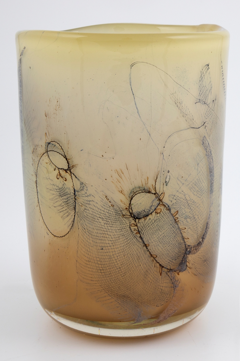 Sylinderformet vase i halvgjennomskinnelig glass. Ujevn munning. Grågrønt underfang som er dekorert med innlagte metalltråder samt partier med finmasket blålig netting. Tidvis brune spetter samt luftbobler i glassmassen. Puntemerke under vasens bunn.