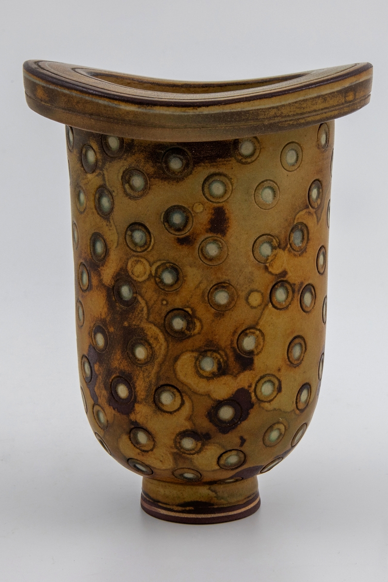 Vase i kraftig steingods. Liten, sirkulær fot. Avlang korpus med rundt tverrsnitt, tykk øvre kant med større ytre diameter enn korpus. Ujevn, flekkete glasur i brunt og olivengrønt. Overflaten utvendig har trykte sirkler der en lysere underglasur kommer frem. Rissede dekorstriper på øvre kant. Et lite uglasert felt nederst på foten med en tynn gulhvit stripe.