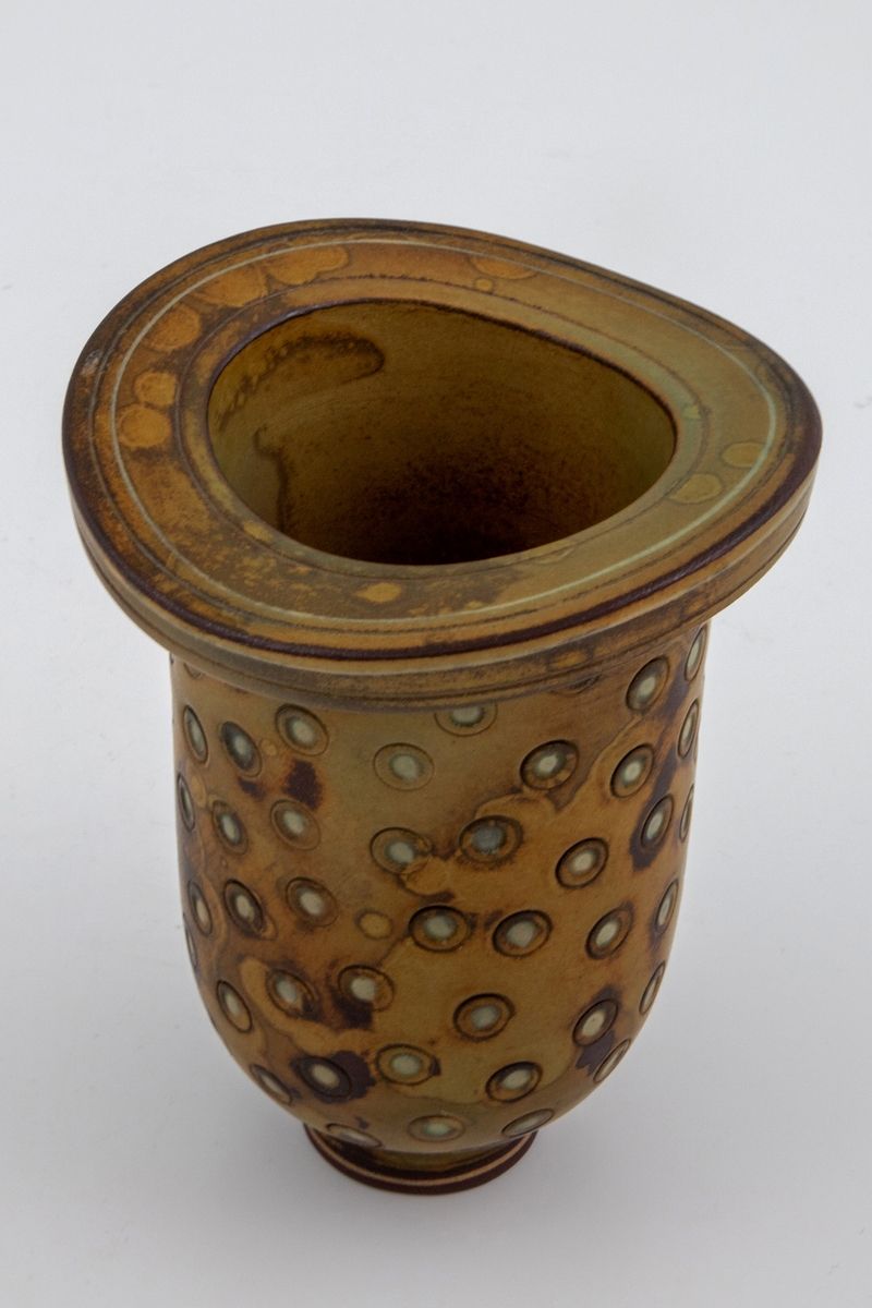 Vase i kraftig steingods. Liten, sirkulær fot. Avlang korpus med rundt tverrsnitt, tykk øvre kant med større ytre diameter enn korpus. Ujevn, flekkete glasur i brunt og olivengrønt. Overflaten utvendig har trykte sirkler der en lysere underglasur kommer frem. Rissede dekorstriper på øvre kant. Et lite uglasert felt nederst på foten med en tynn gulhvit stripe.