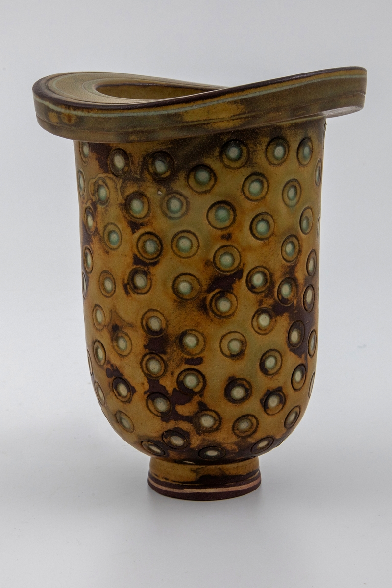 Vase i kraftig steingods. Liten, sirkulær fot. Avlang korpus med rundt tverrsnitt, tykk øvre kant med større ytre diameter enn korpus. Ujevn, flekkete glasur i brunt og olivengrønt. Overflaten utvendig har trykte sirkler der en lysere underglasur kommer frem. Rissede dekorstriper på øvre kant. Et lite uglasert felt nederst på foten med en tynn gulhvit stripe.