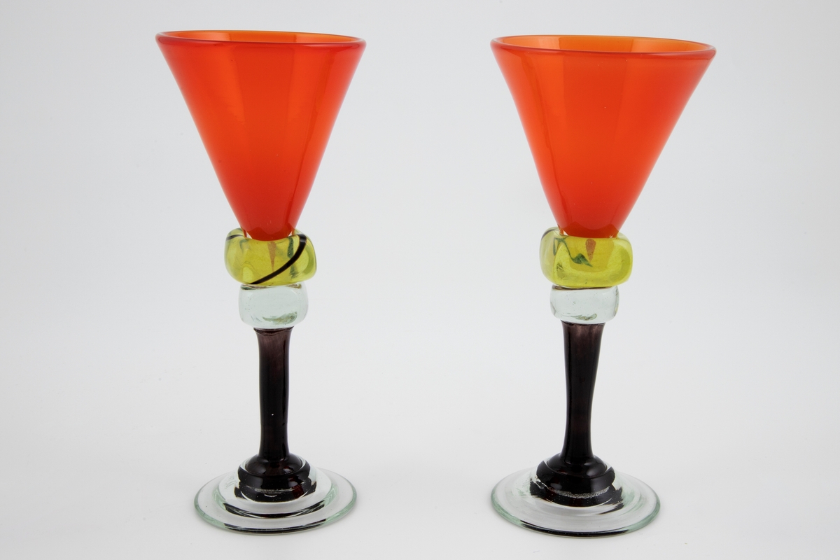 To vinglass i farget glass. Konisk kupa i gjennomskinnelig rødt glass. Sortfarget stett med to terningformede vulster i klart og gult glass, hvorav sistnevnte vulst er dekorert med en bølgende sort linje i spiralform. Sirkulær fot i klart glass, noe hevet, med puntemerke.