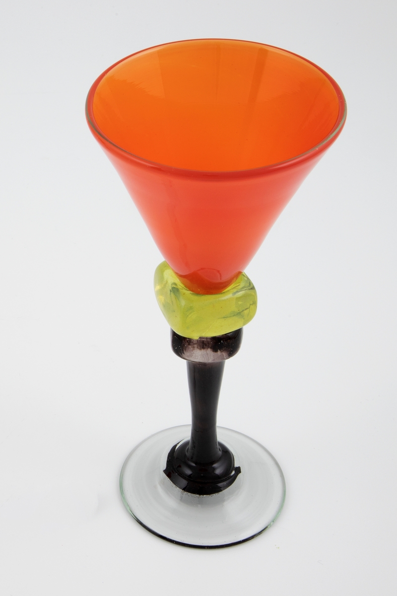 Vinglass i farget glass. Konisk kupa i gjennomskinnelig rødt glass. Sortfarget stett med to terningformede vulster i klart og gult glass, hvorav sistnevnte er dekorert med en bølgende sort linje i spiralform. Sirkulær fot i klart glass, noe hevet, med puntemerke.

