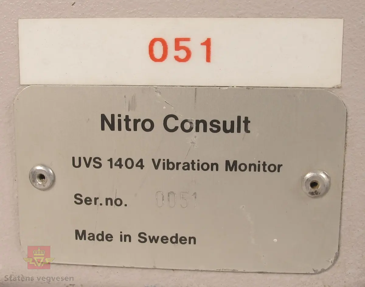 Vibrasjonsmåler UVS 1404. Grålakkert skap i metall som inneholder en vibrasjonsmåler. Bærehåndtak med lær på toppen, fire gummiføtter for stående bruk og fire gummiføtter for liggende bruk, samt brakett for opphenging vertikalt. Lås på skapdør og en lås på nedre del av betjeningspanelet. Utsiden av skapet har påskriftene UVS 1404 Vibration Monitor Nitro Consult fra produsenten, samt klistremerker med påskriftene DYNO Dyno Consult AS Gullaug Postboks 614 3412 LIERSTRANDA Telf: 32 84 12 00 Telefax: 32 22 81 83, TRYKK LOKK INNTIL VED ÅPNING/LUKKING, 051 og 0051. Skiltplate med innskriften Nitro Consult UVS 1404 Vibration Monitor Ser.no. 0051 Made in Sweden på siden av skapet. Vibrasjonsmåleren har påskriften UVS 1404 Vibration Monitor Nitro Consult fra produsenten, og klistremerker med påskriftene DYNO Dyno Consult AS Gullaug Postboks 614 3412 LIERSTRANDA Telefon: 32 84 12 00 Telefax: 32 22 81 83 og AVLESNING SE INNSIDEN AV LOKK. Vibrasjonsmåleren har to betjeningspanel, det ene under låsbart lokk og avlesningsvinduer. På siden av skapet er det tilkoblingsmuligheter for kabler. 2 stk 6 volts batterier med påskriften DURACELL PROCELL PROFFESIONAL ALKALINE BATTERY PC908 6 VOLT BEST IF INSTALLED BY: JAN 2002.