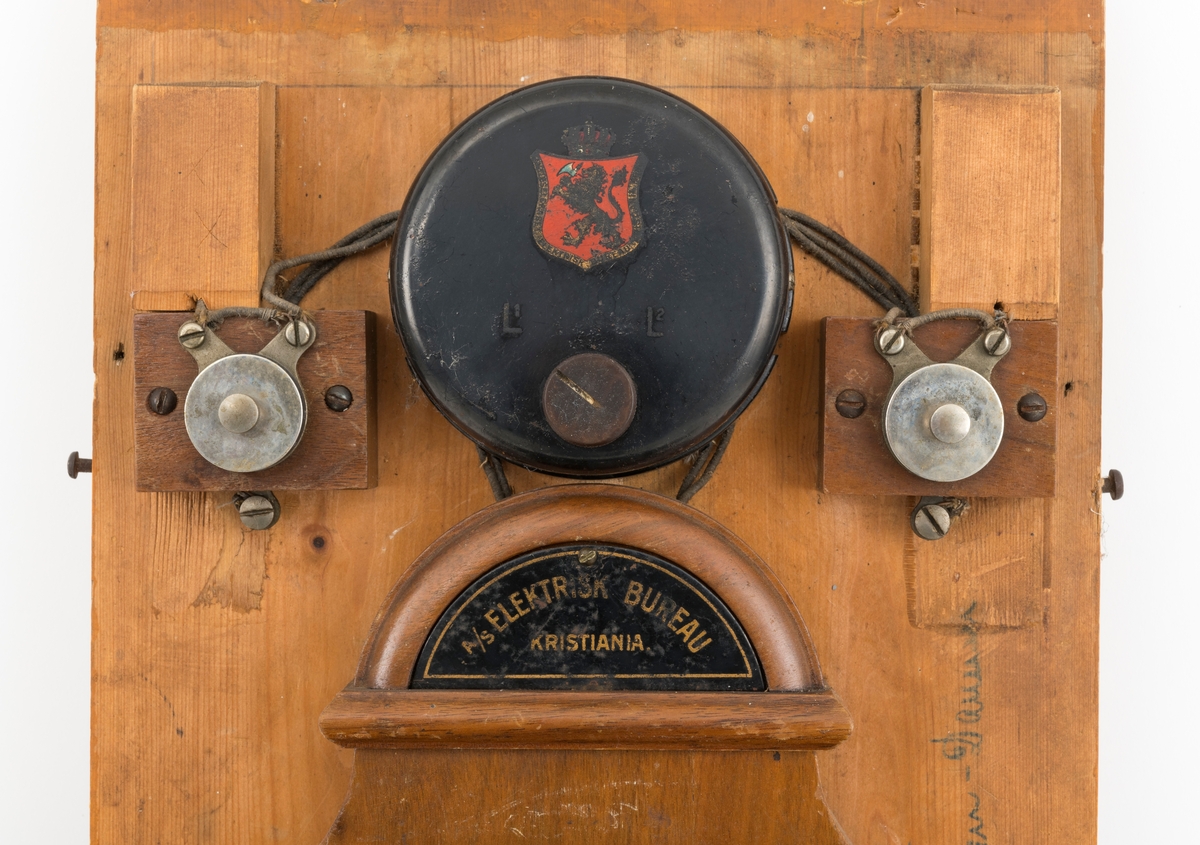 Sveivetelefon, antakelig av type som ble brukt omkring 1. verdenskrig.  Telefonen er montert på ei 1,6 centimeter tjukk treplate.  Apparatet består av ei "kasse" av tre og jernblikk, alt lakkert i edeltreimiterende brunfarge.  I denne delen av apparatet inngår et skrått lokk, en liten skriveklaff, som antakelig skjermer underliggende mekanikk og muligens også batterier.  Ellers har apparatet en forkrommet holder for telefonrøret, som også er forkrommet, men har bakelittkomponenter for håndgrep (midten), øreanlegg og taletrakt.  Apparatet har to omvendt skålformete forkrommete bjeller bak rørholderen, som antakelig gav fra seg klemt når det kom henvendelser per telefon.  Øverst på apparatet er det et slags halvsirkelformet "hode" med informasjon om produsenten.  Her er det også et rødt skjold med en løvefigur med krone over.  Dene logoen finnes også på en svartlakkert, sylindrisk boks på apparatet.  Fra denne boksen er det ledningsforbindelse til stålskiver, som er fastskrudd på begge sider av apparatet. Plata telefonen er montert på har blyantpåskriften «Stor-En-Dammen». Dette antas å være en god indikasjon på at dette apparatet har vært montert i koia ved den nevnte dammen i et av Osensjøens sidevassdrag. Her ble telefonen brukt når fløterne i nedenforliggende deler av vassdraget ønsket å gjøre avtaler som regulerte vanntilførselen i elveløpet på en måte som var optimal ned henblikk på å få tømmeret til å flyte mot Osensjøen.