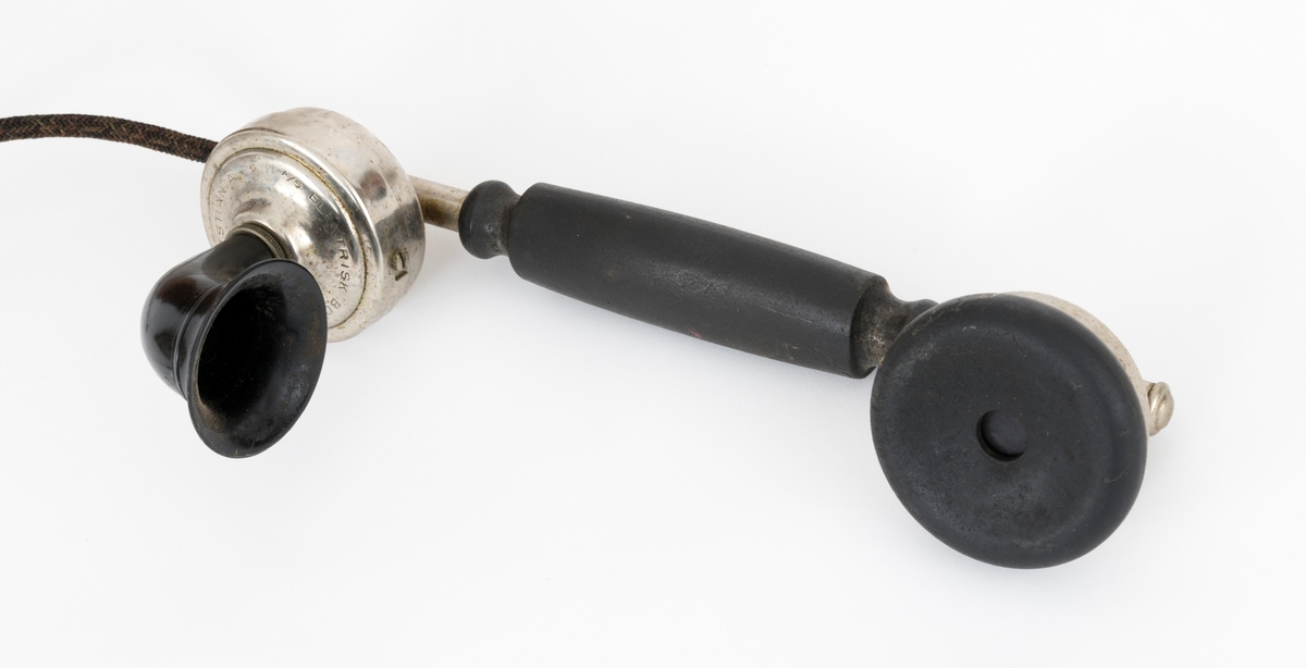 Sveivetelefon, antakelig av type som ble brukt omkring 1. verdenskrig.  Telefonen er montert på ei 1,6 centimeter tjukk treplate.  Apparatet består av ei "kasse" av tre og jernblikk, alt lakkert i edeltreimiterende brunfarge.  I denne delen av apparatet inngår et skrått lokk, en liten skriveklaff, som antakelig skjermer underliggende mekanikk og muligens også batterier.  Ellers har apparatet en forkrommet holder for telefonrøret, som også er forkrommet, men har bakelittkomponenter for håndgrep (midten), øreanlegg og taletrakt.  Apparatet har to omvendt skålformete forkrommete bjeller bak rørholderen, som antakelig gav fra seg klemt når det kom henvendelser per telefon.  Øverst på apparatet er det et slags halvsirkelformet "hode" med informasjon om produsenten.  Her er det også et rødt skjold med en løvefigur med krone over.  Dene logoen finnes også på en svartlakkert, sylindrisk boks på apparatet.  Fra denne boksen er det ledningsforbindelse til stålskiver, som er fastskrudd på begge sider av apparatet. Plata telefonen er montert på har blyantpåskriften «Stor-En-Dammen». Dette antas å være en god indikasjon på at dette apparatet har vært montert i koia ved den nevnte dammen i et av Osensjøens sidevassdrag. Her ble telefonen brukt når fløterne i nedenforliggende deler av vassdraget ønsket å gjøre avtaler som regulerte vanntilførselen i elveløpet på en måte som var optimal ned henblikk på å få tømmeret til å flyte mot Osensjøen.