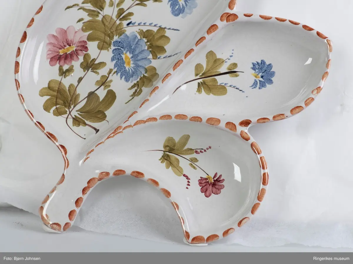 Keramikkfat formet som et blad, med ett stort og to små rom. Dekorert med håndmalte blomster. Stilken på bladet er knekt av og mangler. Antakelig produsert av Arol keramikk i Halden.