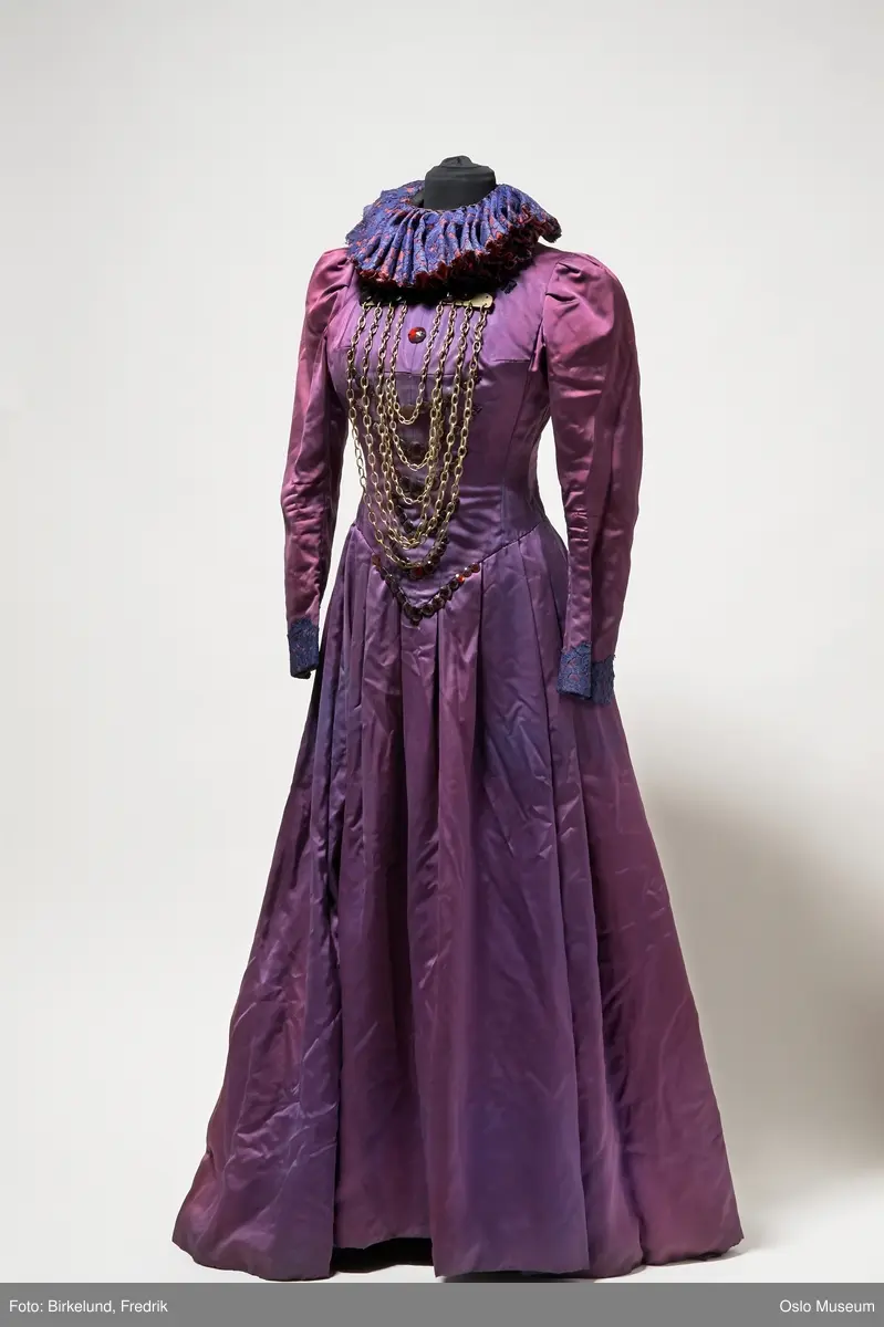Lilla kjole i renessansestil. Innebygget korsett. Lange ermer med blå blondedekor ved håndleddet. Fyldig krave rundt halsen i rødt tekstil trukket med blå blonde. Kjolen lukkes med hekter i ryggen. Dekorert med røde steiner og gullfarget kjetting på frontpartiet av kjolen.