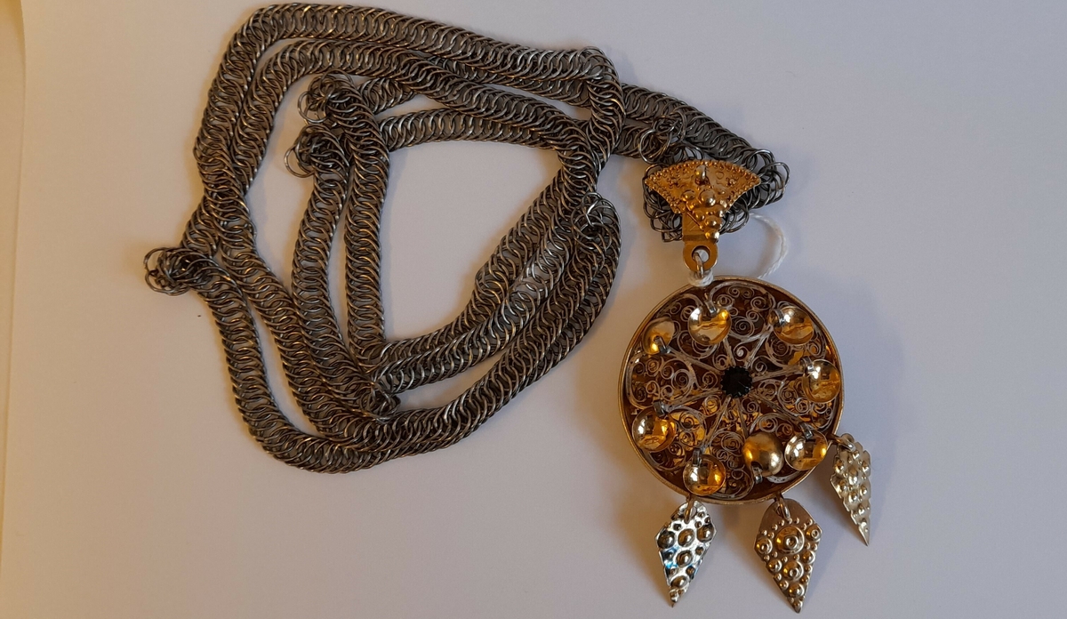Skålforma botn, filigranslok med raud stein i midten og åtte lauv i krans rundt. I underkant tre prismeforma lauv. Kjedefestet er forma som i krune med prismelauv. Lang flatkjede.

SUM. tilvekstprotokoll 1955:
Sylgje m. kjede. Gylt sølv, diam 5,5. Form konkav med konvekst filigransverk, raud stein og lauv. Stempla N, Bergen 1825.