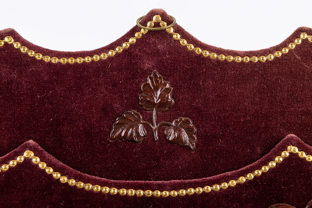 Väska att hänga på väggen, i papp överklädd med rödlila sammet och garnerad med pressade pappmotiv i brunt och guld. I mitten broderat mönster i korsstygma på vit stramalj.