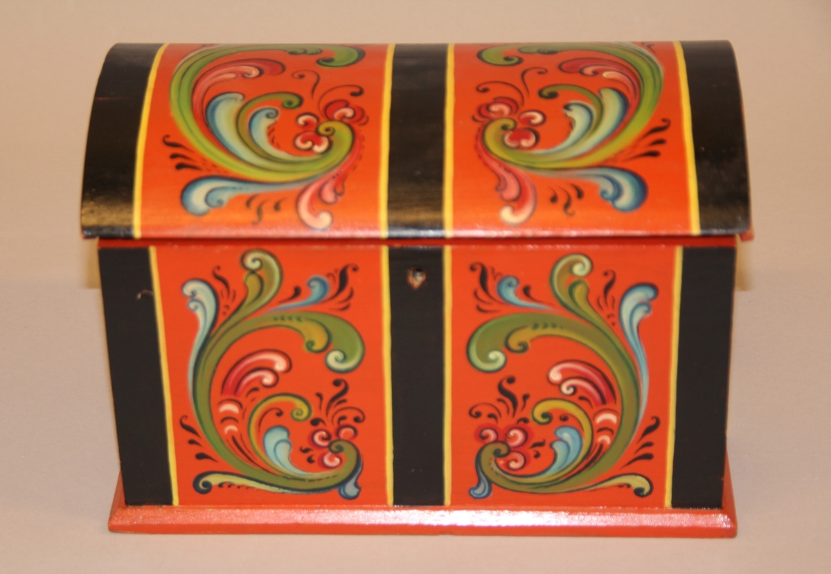 Lita kiste med kuva lok. Rosemåla med tydelege c-krullar. Einsfarga inni, eit lite rom med lok på den eine sida inne i kista.