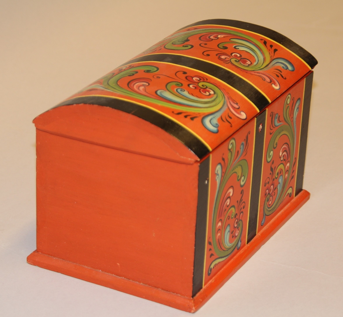 Lita kiste med kuva lok. Rosemåla med tydelege c-krullar. Einsfarga inni, eit lite rom med lok på den eine sida inne i kista.