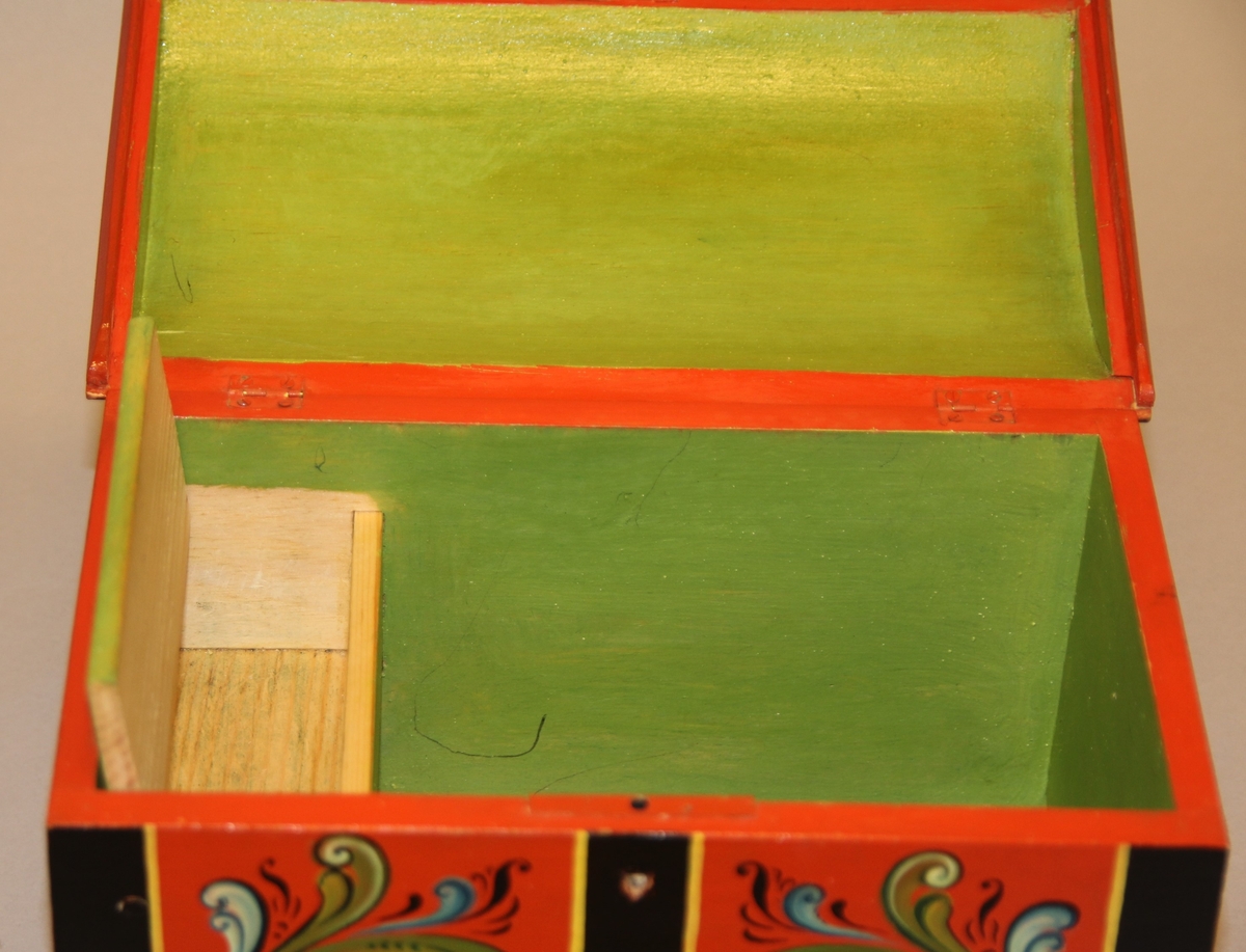 Lita kiste med kuva lok. Rosemåla med tydelege c-krullar. Einsfarga inni, eit lite rom med lok på den eine sida inne i kista.