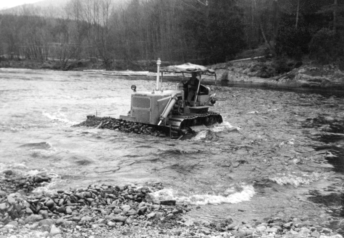 Bulldoser, fotografert i samband med "elverenskingsarbeid" i Skiensvassdraget, antakelig i 1950-åra.  Skiensvassdragets Fellesfløtningsforening leide bulldoser med sikte på fjerning av stein- og grusører i elveløpet i Bøelva i 1953.  På denne måten håpte organisasjonen å kunne redusere problemet med at det la seg hauger eller vaser på slike grunner.  Elverenskingsoppdraget ble lagt ut på anbud, og det var Tele Maskinsentral ved Halvor K. Skjøllaug som fikk oppdraget.  Fotografiet viser beltekjøretøyet som skyver stein- og grusmasser fra elveløpet mot den ene elvebredden.  I det nedenforleggende elveløpet, hvor maskinen har gått, framstår elveløpet som forholdsvis rett og stilleflytende. Samme sesong som den avbildete bulldoseren arbeidet i Bøelva ble det også gjort liknende elverenskingsarbeid i Bygdaråi i Seljord (i nærheten av Lislandbrua), i Flatdal (antakelig Åmotsdalselva ovenfor Flatdalsvatnet) og i Hjartdøla i Hjartdal.  I slutten av 1950-åra påpekte Bø og Nes jakt og fiskelag at Fellesfløtningsforeningen ved denne formen for elverensk kjørte fiskeyngel på land og ødela gyteplassene.  Sportsfiskernes interesseorganisasjon krevde at den ødelagte yngelen ble ersttattet av settefisk, noe Skiensvassdragets Fellesfløtningsforening etter hvert aksepterte. 