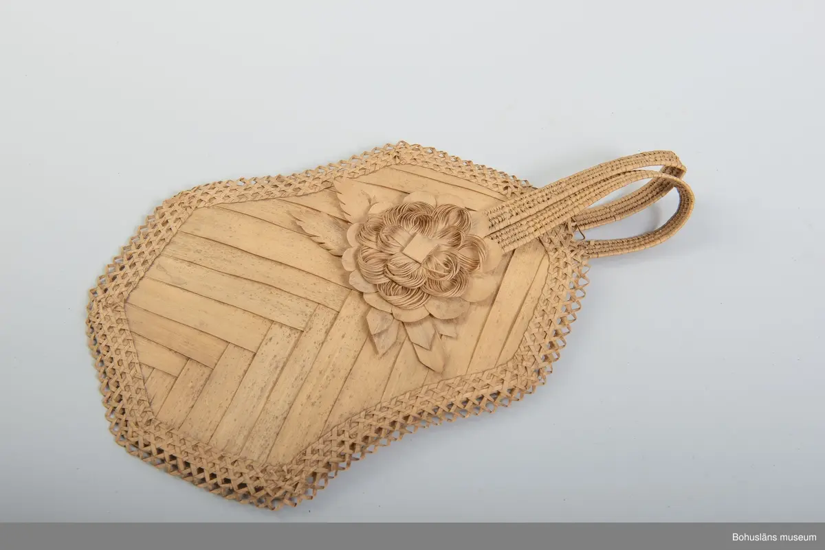 Ur handskrivna katalogen 1957-1958:
Solfjäder av trä, Amerika
Mått: 30 x 18 cm; av bast och tunna träfibrer; en mångsid. platta m. runda hörn, jämte handt. Hel.

Ur handskriven katalog över Uddevalla - Musei Historiska - samlingar: Utländska föremål = De etnografiska föremålen upprättad  år 1916 av Knut Adrian Andersson, Intendent från 1 Oktober 1915:
Solfjäder (Fläkt) av trä (     ), gjord av infödingar å St Barthélèmy 1840talet. Skänkt av fröken Anna Craelius i Uddevalla.

Troligen gjort av strå av någon sorts palmblad.