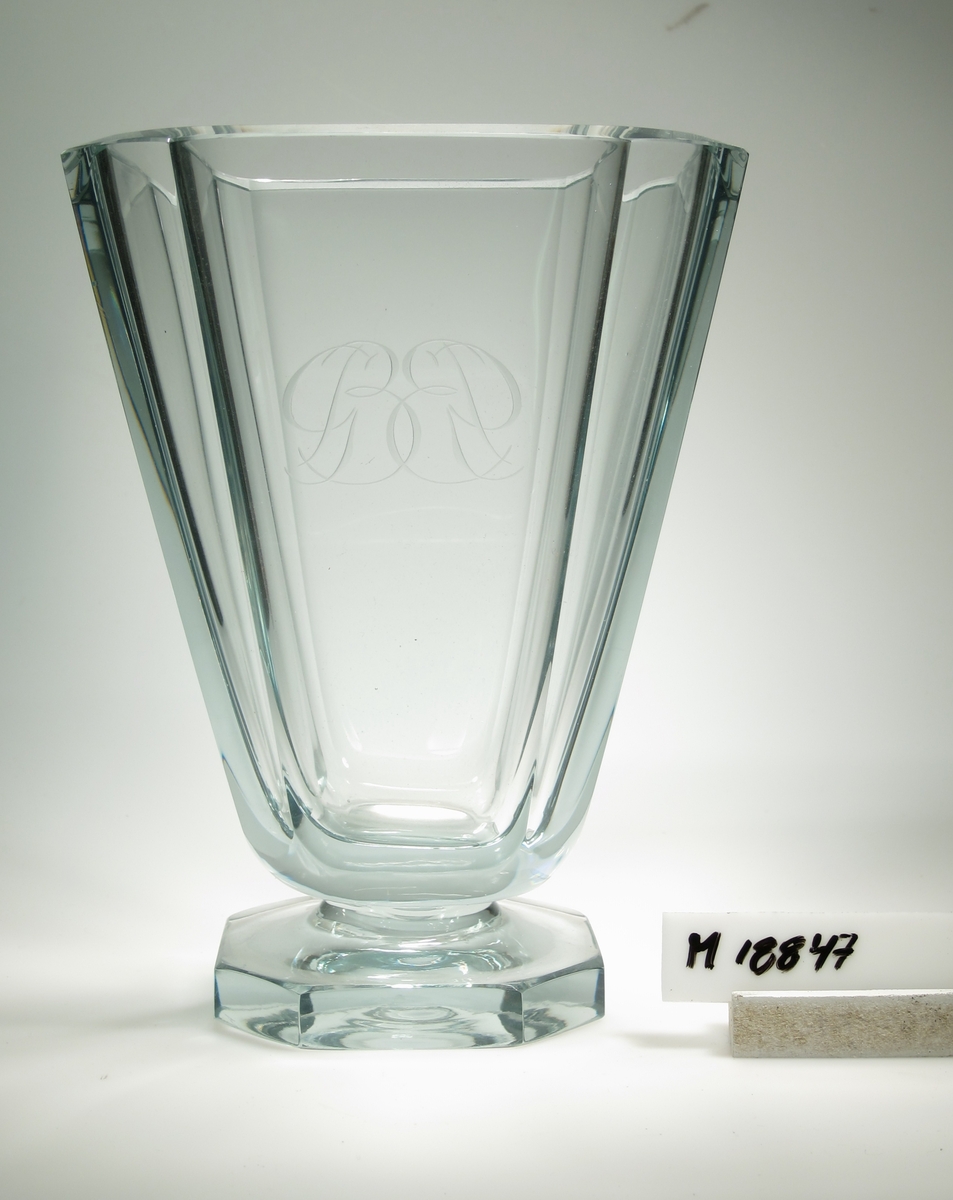Vas på fot designad av Gerda Strömberg. Grönblåtonat glas, tjockt i godset, facettslipade kanter på vasen och slipad 8-kantig fot. Graverat monogram "GB". Artikelnummer B485.