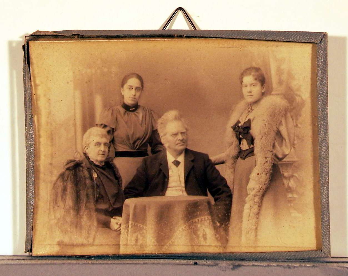 Atelierfotografi; bakerst står to jevngamle unge damer, den ene med lang pelsstola hengende ned langs med skjørtet og en stor mørk sløyfe bundet midt for brystet; den andre unge dame mer enkelt kledd i lys lang kjole med mørkt bånd rundt taljen og halsen isatt enkel brosje. Inn til et lite rundt bord sitter en dame med pelskape til høyre, og i midten en mann med henderne i siden, nesebriller, mørk jakke og slips, hvit skjorte og vest. Teppebakgrunnen viser antikke bygningsfragmenter. De tre damene ser direkte på betrakteren, mens mannen ser skrått til venstre.