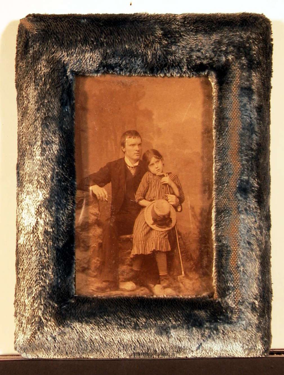 Atelierfotografi av voksen ung mann sittende med en stor pike på kneet og støttende seg til en lang stokk med ene hånd og holdende en sigarett i høyre hånd. Piken har også tak i stokken og i høyre hand holder hun en stor stråhatt med bredt bånd og sløyfe.
Bakgrunnen viser et fjelllandskap.