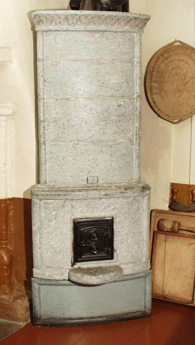 Klebersteinsovn med utskåret bord på toppstykket. Ovnsdøren er i smijern. Ovnen har et fotstykke i gråmalt tre.