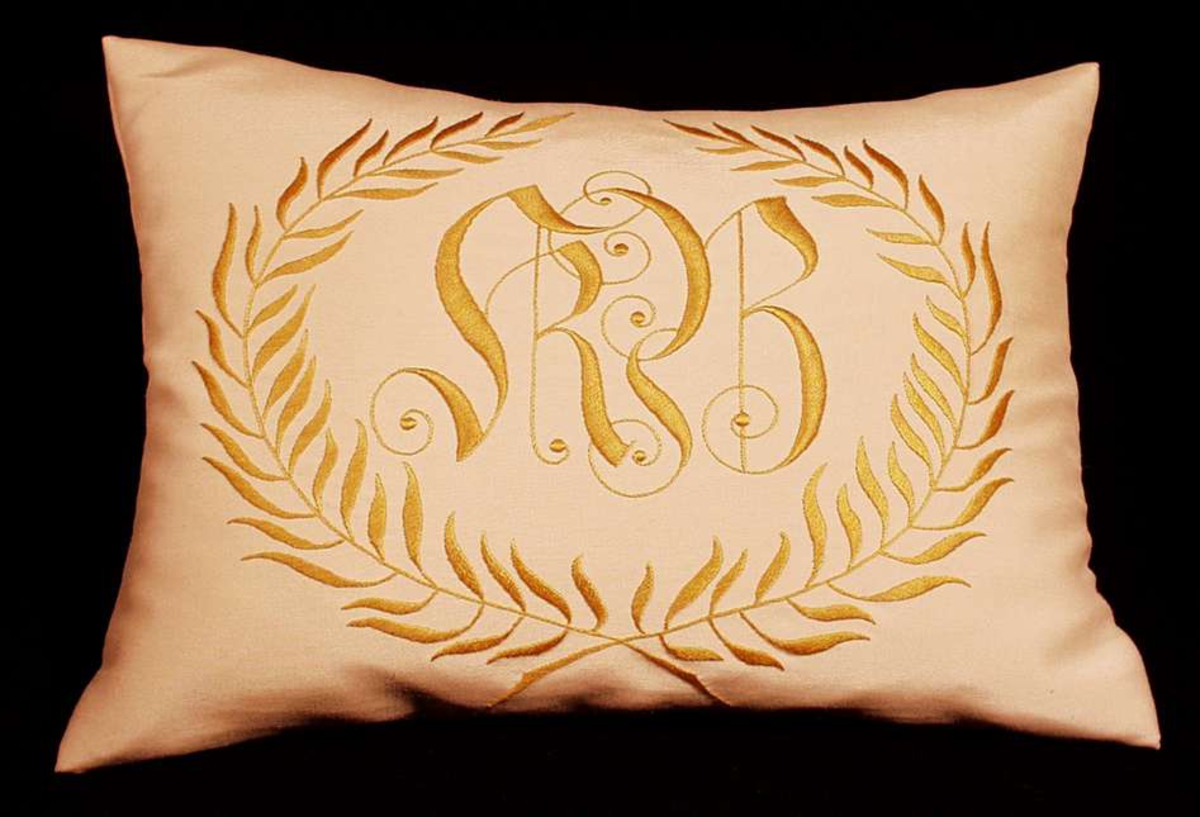 Monogram i laubærkrans brodert på silkerips.  De to laurbærgrenene er brodert med stilkesting og plattsøm. Monogrammet er brodert med stilkesting og tykksøm.
Mangler trykknapper.