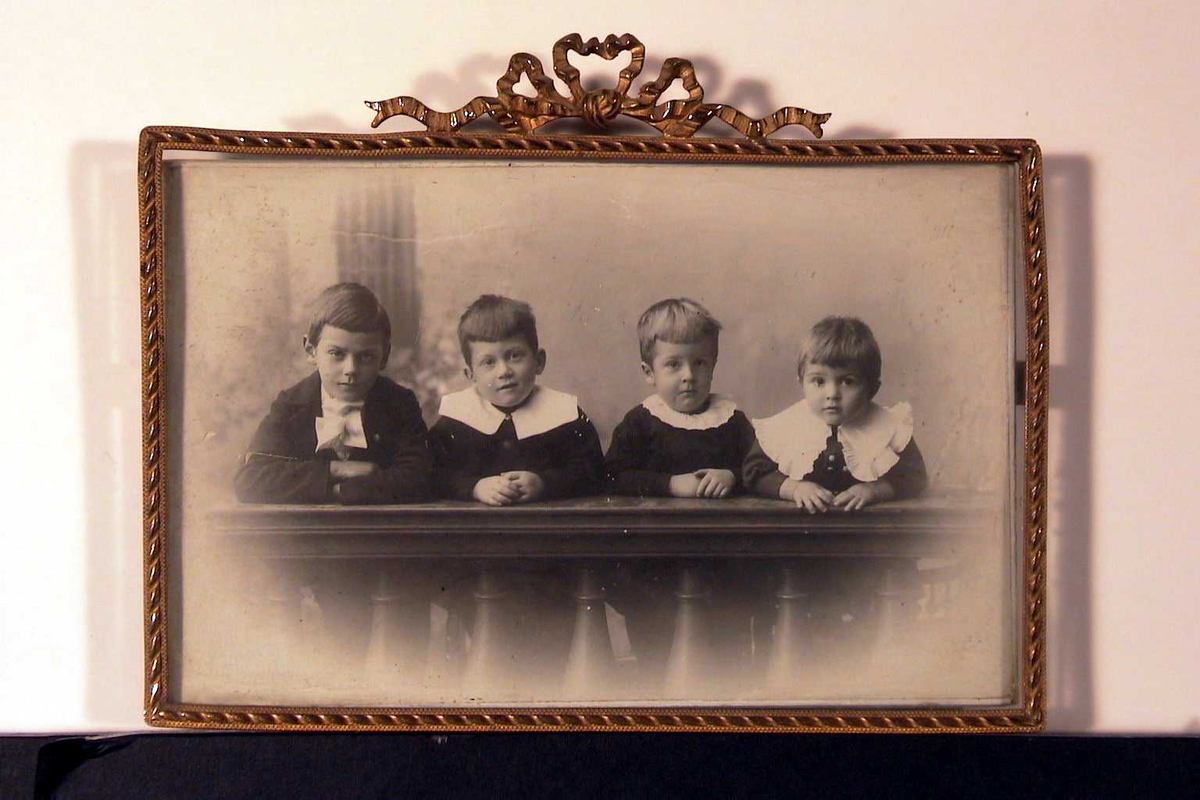 Gruppeportrettt. Fire gutter bak ballustrade-arrangement. Alle i svarte dresser, de yngste med løse, hvite krager, den eldste med hvit sløfe. Alle lener seg mot gelenderet i fotoatelieet.