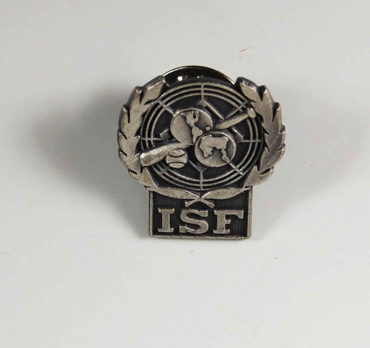 Sølv- og gråfarget pins med logo for ISF. I logoen inngår det en ball og et balltre. Logoen utgjør pinsens form.