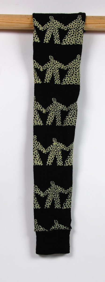 Sort og hvit knestrømpe til vettedrakt. Det ser ut til at knestrømpen er laget ved å klippe bena av en stilongs. Det er ikke noen søm på strømpens øvre kant, og kanten er også ujevn. Menneskefigurer som holder hver andre i hendene utgjør dekoren på strømpen.