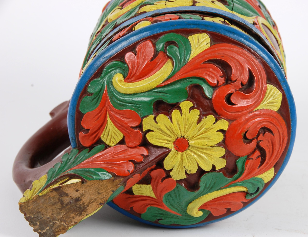 Kanne av staver. Lokk med gule, grønne og røde blader, blåmalt kant. Korpus med en blåmalt metallgjord oppe og nede, gule, grønne og røde akantusblader, brunmalt hjerteformet felt med innskriften 1865. Brunmalt hank. Tre røde og gule føtter.