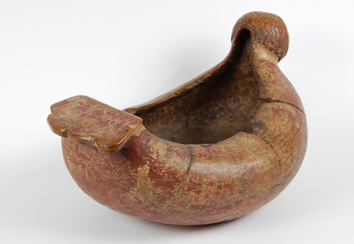 Trøys av bjørk. Oval form. Utskåret mannshode med munnåpning. Håndtak med hakkformete utskjæringer og innskrift. Svarte bekklumper i bunnen.